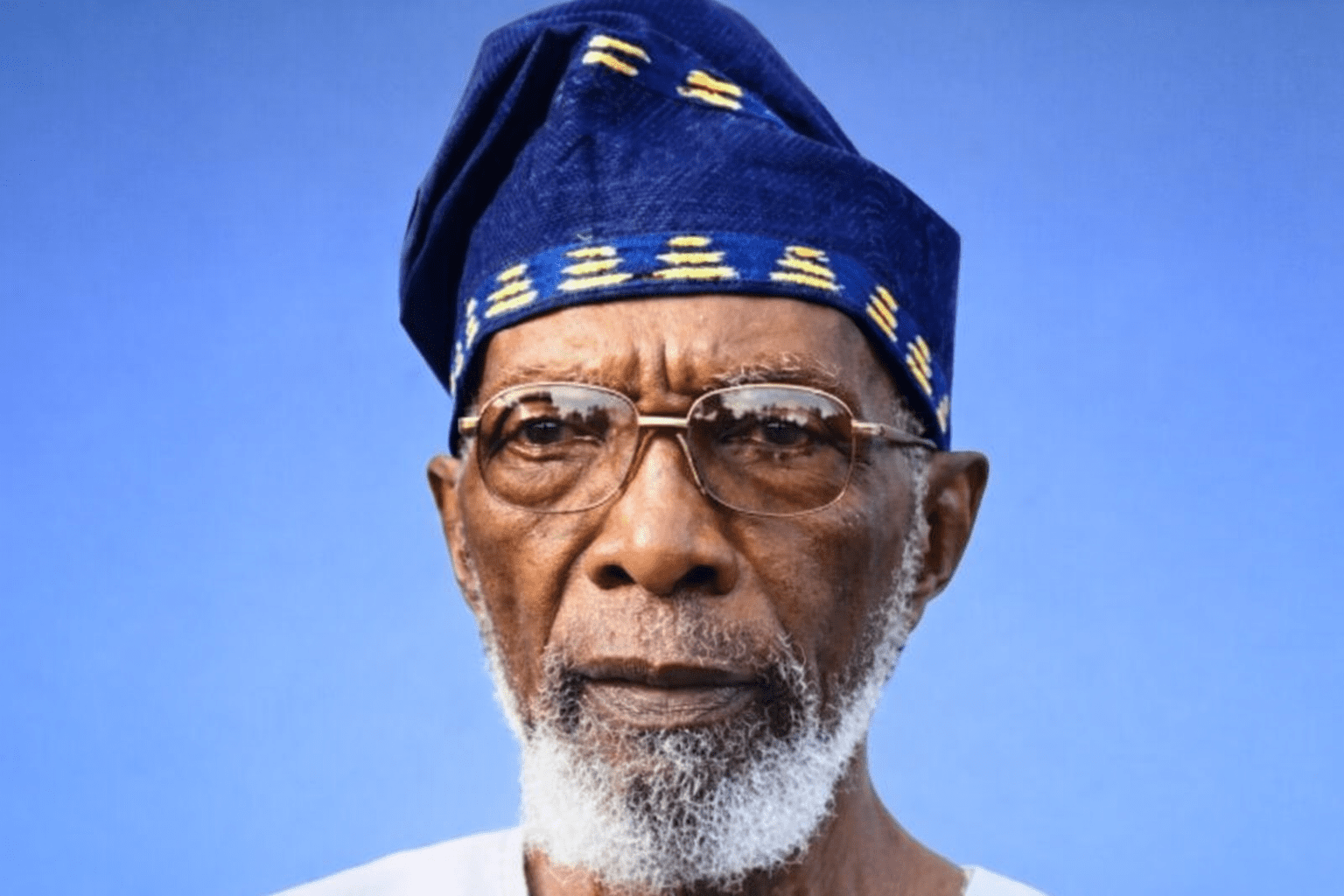 Portrait of Dr Segun Osoba wearing a navy blue Yoruba cap and white traditional attire, looking composed against a soft blue background.