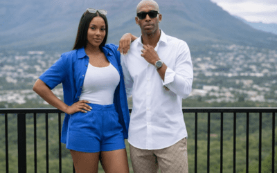 Stylish couple standing on a balcony with a mountain backdrop, the woman in a blue outfit and the man in a white shirt.