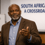 A close‑up of Professor Wole speaking on stage in a South African university lecture hall, gesturing as he explains the theme “South Africa at a Crossroads,” with the word “WHY?” visible on a whiteboard behind him.