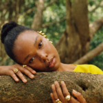 Aya resting her head and hands on a textured tree branch in a tranquil forest, wearing gold rings and ear cuffs that glint softly in the filtered sunlight. Her bright yellow top contrasts with the earthy tones of the bark and foliage, creating a serene portrait of calm and connection with nature.