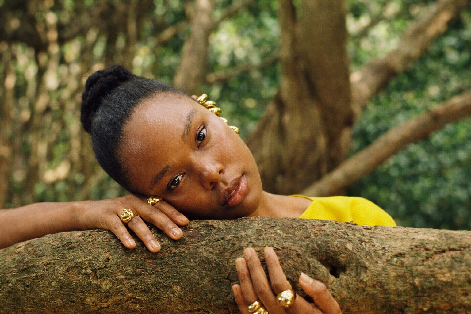 Aya resting her head and hands on a textured tree branch in a tranquil forest, wearing gold rings and ear cuffs that glint softly in the filtered sunlight. Her bright yellow top contrasts with the earthy tones of the bark and foliage, creating a serene portrait of calm and connection with nature.