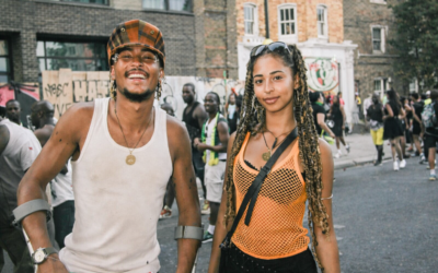 Two people standing together at a Caribbean cultural event in a British city, wearing bright clothing and Caribbean flags, with festival goers and urban buildings behind them.