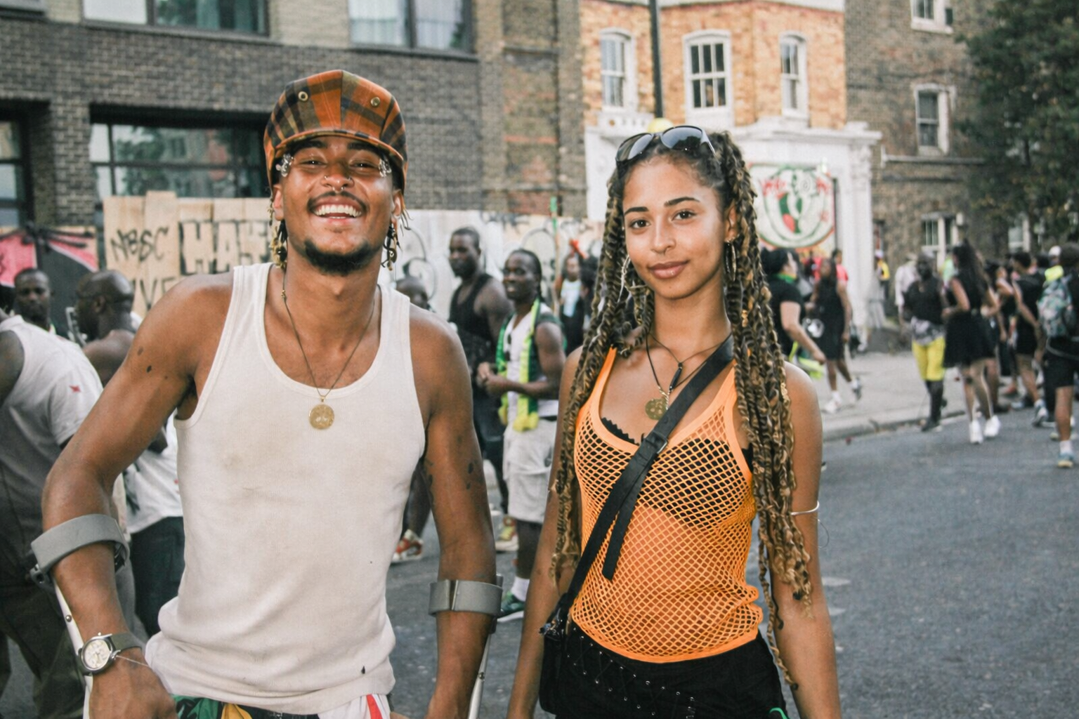 Two people standing together at a Caribbean cultural event in a British city, wearing bright clothing and Caribbean flags, with festival goers and urban buildings behind them.