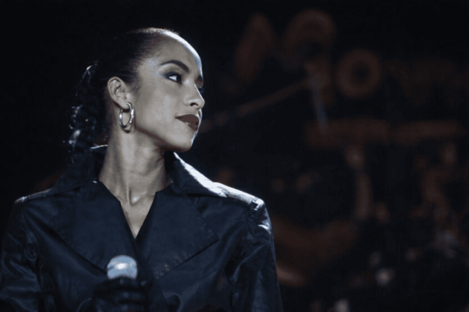 Sade Adu performing live on stage, wearing a dark satin outfit and silver hoop earrings, holding a microphone under soft stage lighting.
