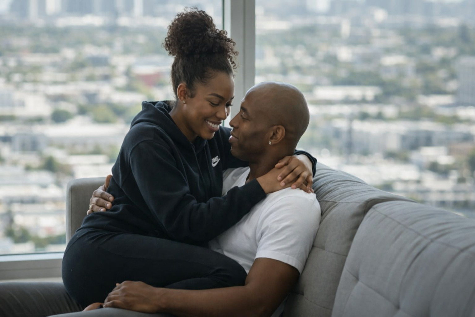 Sola and Peter sit closely on a light grey couch in their UK living room, with soft daylight streaming through a large window behind them. The scene captures warmth and affection as they share a relaxed, intimate moment.