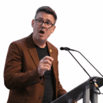 Andy Burnham, the UK Prime Minister, wearing glasses and a brown jacket, speaking at a podium.