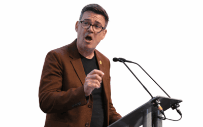 Andy Burnham, the UK Prime Minister, wearing glasses and a brown jacket, speaking at a podium.