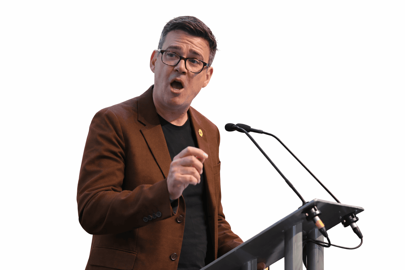 Andy Burnham, the UK Prime Minister, wearing glasses and a brown jacket, speaking at a podium.