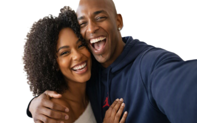 Sola and Peter stand close together in their London living room, smiling brightly at the camera. Sola wears a light top with curly hair framing her face, while Peter wears a dark blue hoodie with a red logo. The plain white background highlights their joyful expressions and closeness.