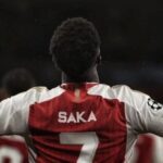 Bukayo Saka, wearing his red and white Arsenal jersey with the number 7, raises both arms with index fingers pointing upward during a UEFA Champions League game.