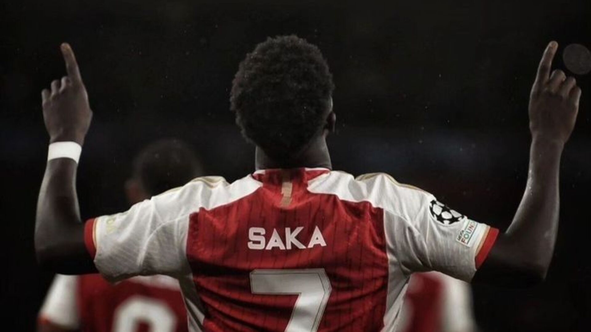 Bukayo Saka, wearing his red and white Arsenal jersey with the number 7, raises both arms with index fingers pointing upward during a UEFA Champions League game.