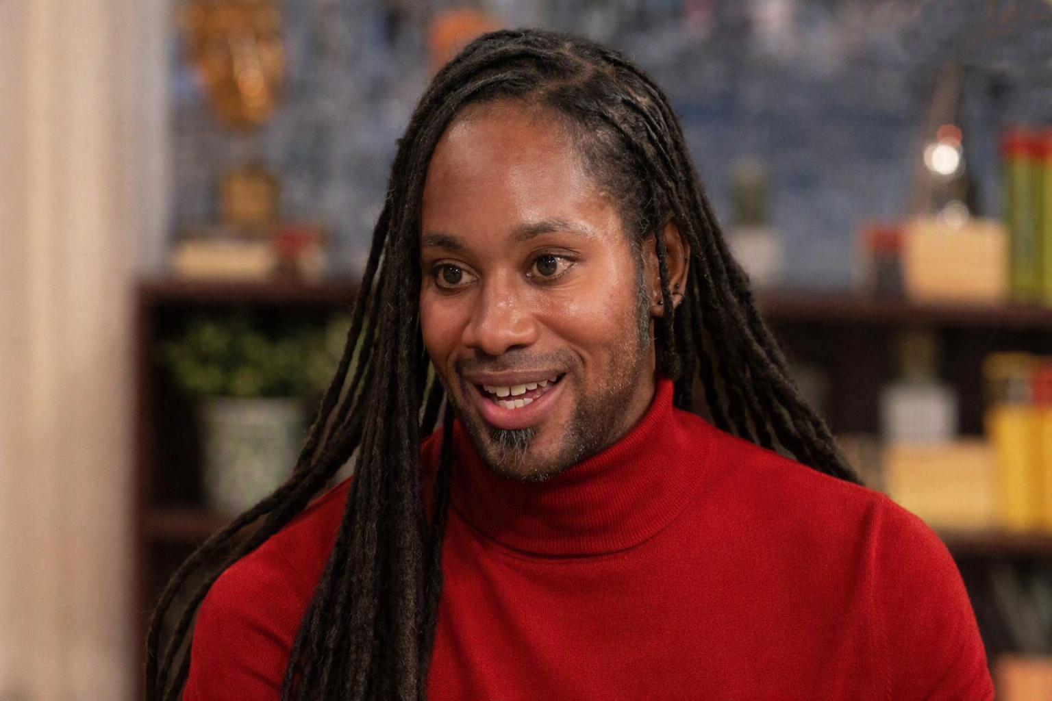 Jason Arday smiling in a red turtleneck sweater, seated indoors with books and plants in the background.