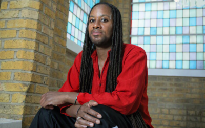 Jason Arday seated indoors near stained‑glass windows wearing a red shirt and black trousers with white stripes.