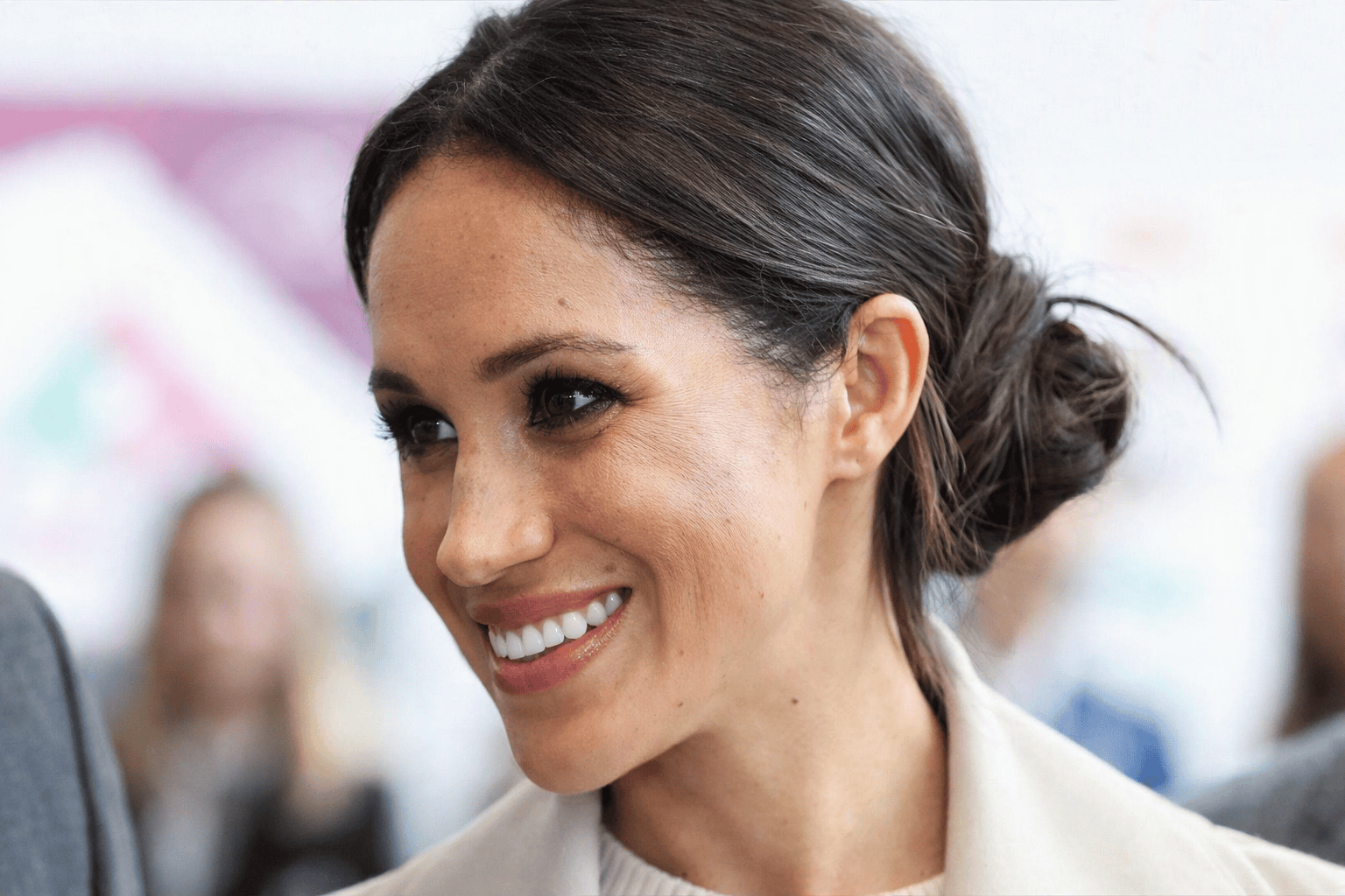 Meghan Markle smiling and waving at an outdoor public event, surrounded by attendees wearing lanyards.