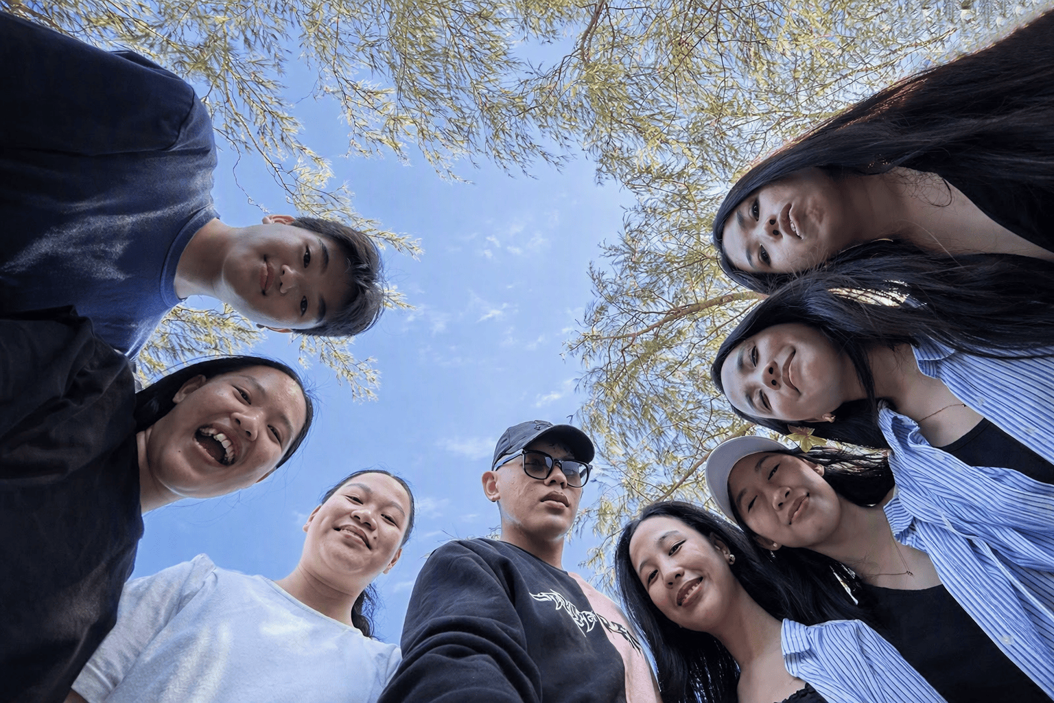 Nine young friends standing in a circle on a London street, viewed from below with the sky and tree branches visible above.