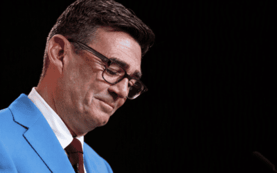 Andy Burnham speaking at a political event, wearing a sky blue suit and burgundy tie against a dark background