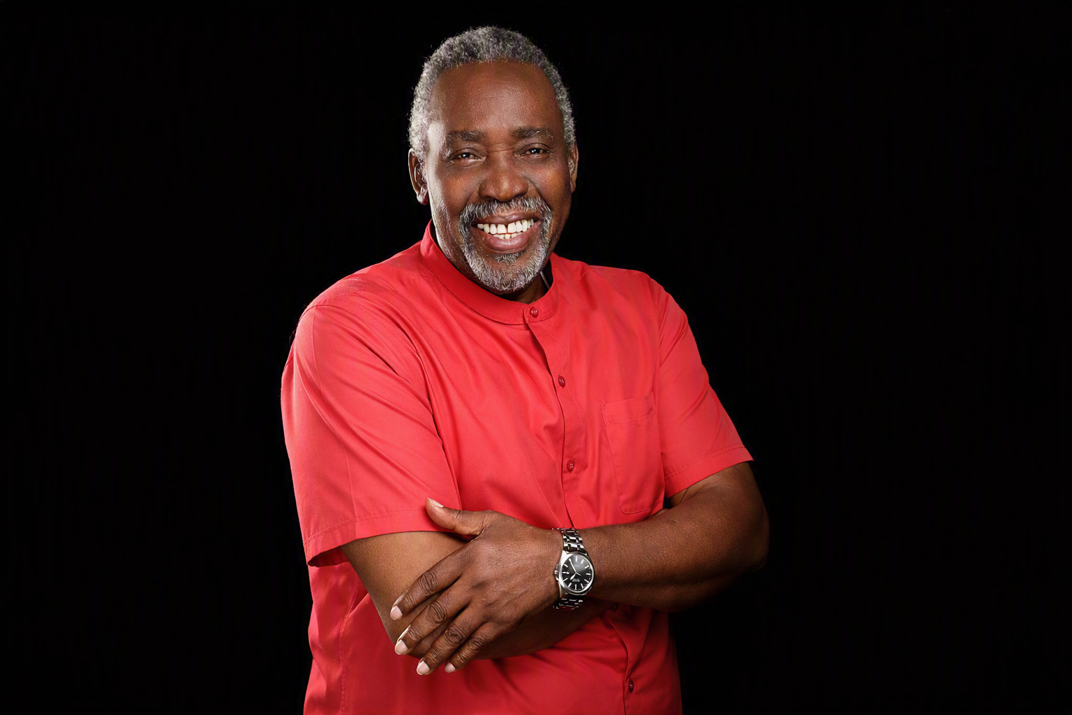 Portrait of Olu Jacobs smiling in a light red shirt against a dark background, symbolising warmth and legacy.