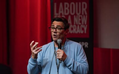 A speaker holding a microphone in front of a red backdrop reading “Labour for a New Democracy,” addressing an audience during a political discussion.