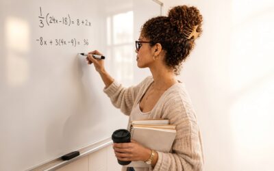 A young teacher in a UK classroom explaining algebraic equations on a whiteboard while holding a notebook and coffee cup.
