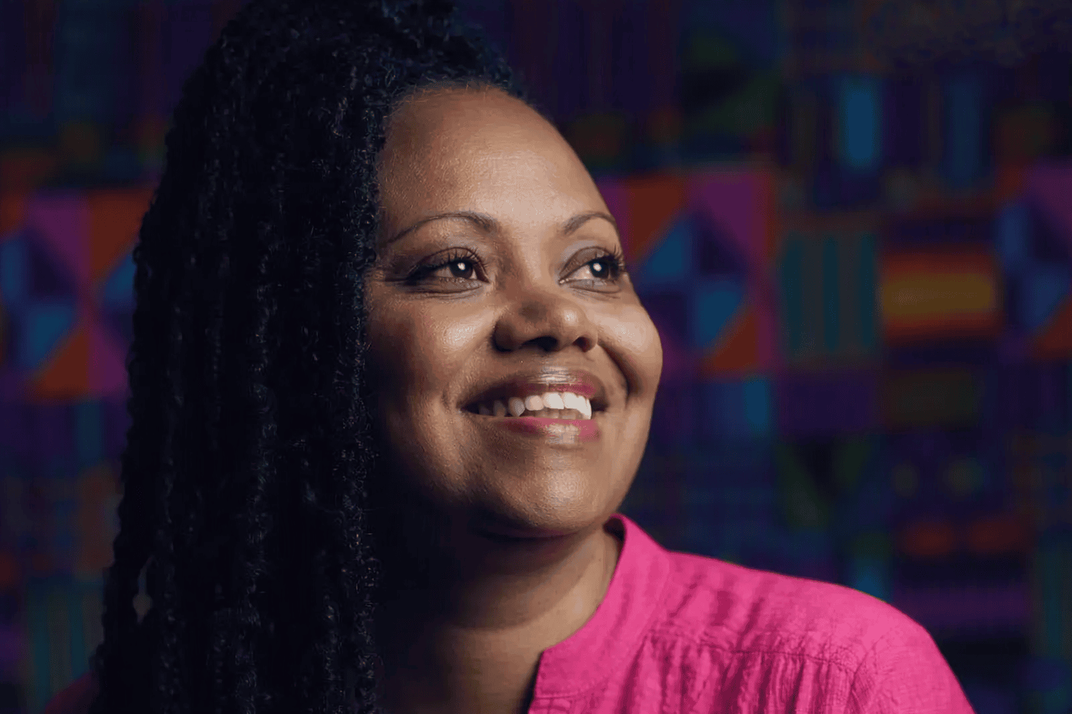 Portrait of Adriana Barbosa smiling in a pink outfit against a colourful geometric background.