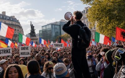 Young man with megaphone addressing a large diverse crowd of protesters in a European city square, holding signs reading “STRIKE”, “GRÈVE” and “HUELGA”, with French, Italian, Spanish and Albanian flags waving under sunny autumn light.